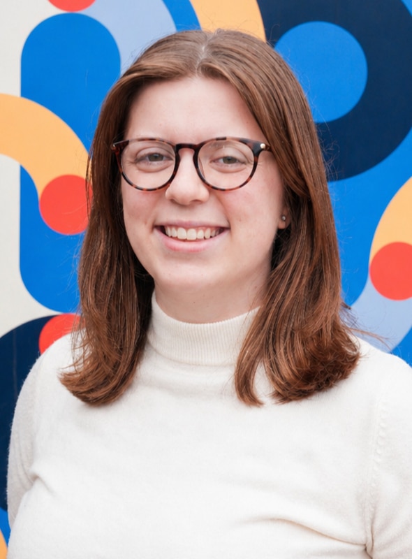 A woman with straight brown hair and glasses smiles, wearing a white turtleneck sweater. The background is a colorful abstract mural with blue, red, and yellow shapes.