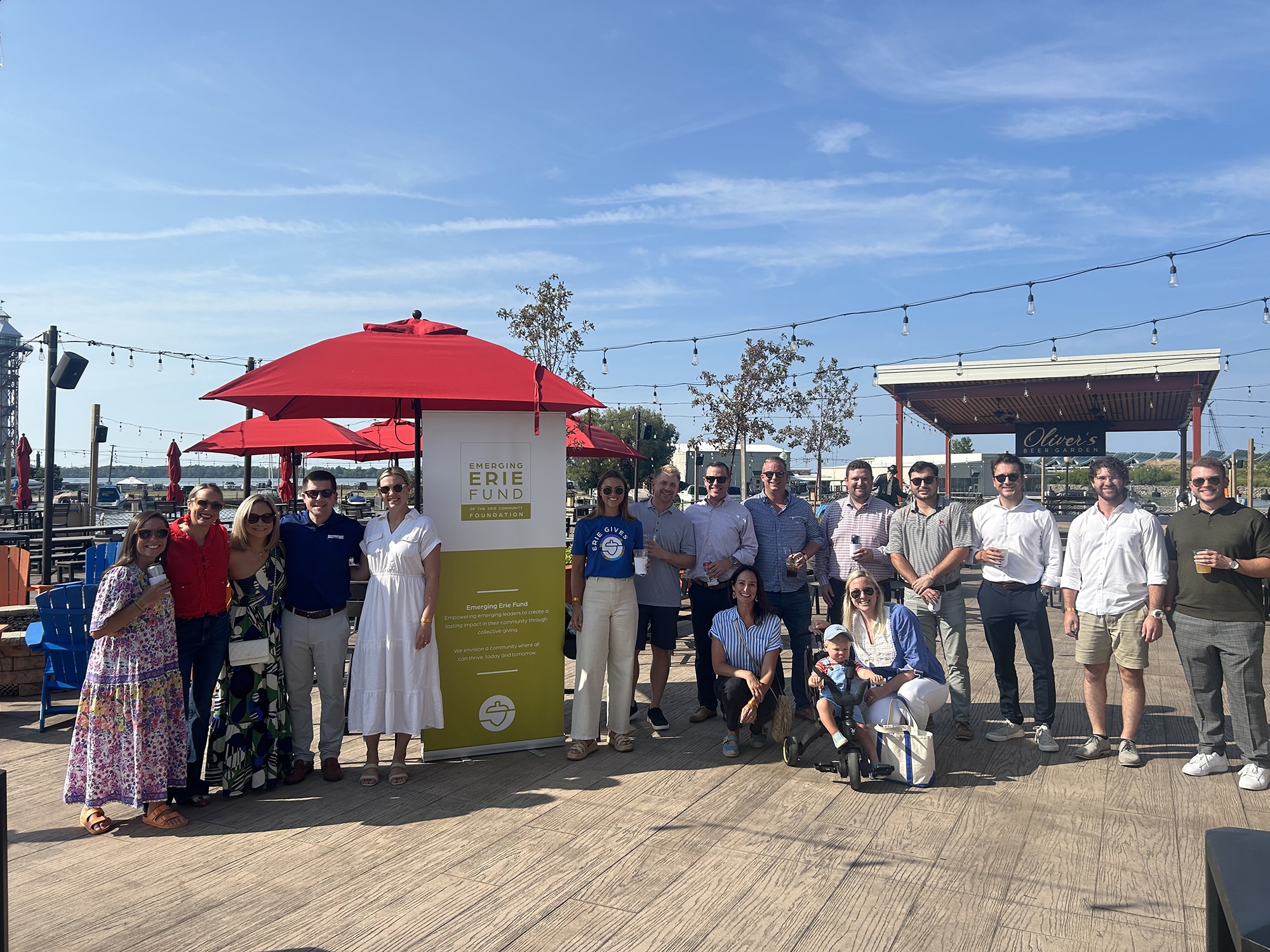 A group of people stand and smile together outdoors on a sunny day by a yellow sign for the Erie Fund, with red umbrellas and string lights visible in the background.
