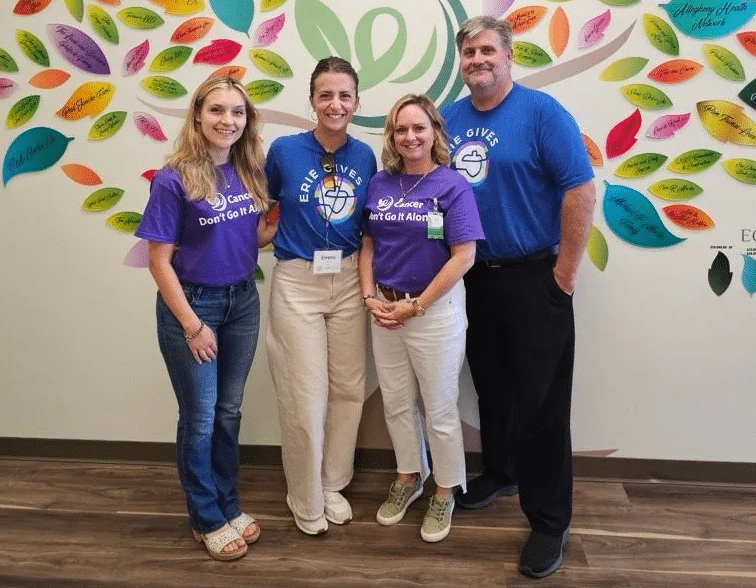 Four people stand smiling in front of a colorful leaf-covered mural. Three wear “Cancer: Don’t Go It Alone” t-shirts in purple or blue, and one wears a light blue shirt. They stand together on a wood floor indoors.