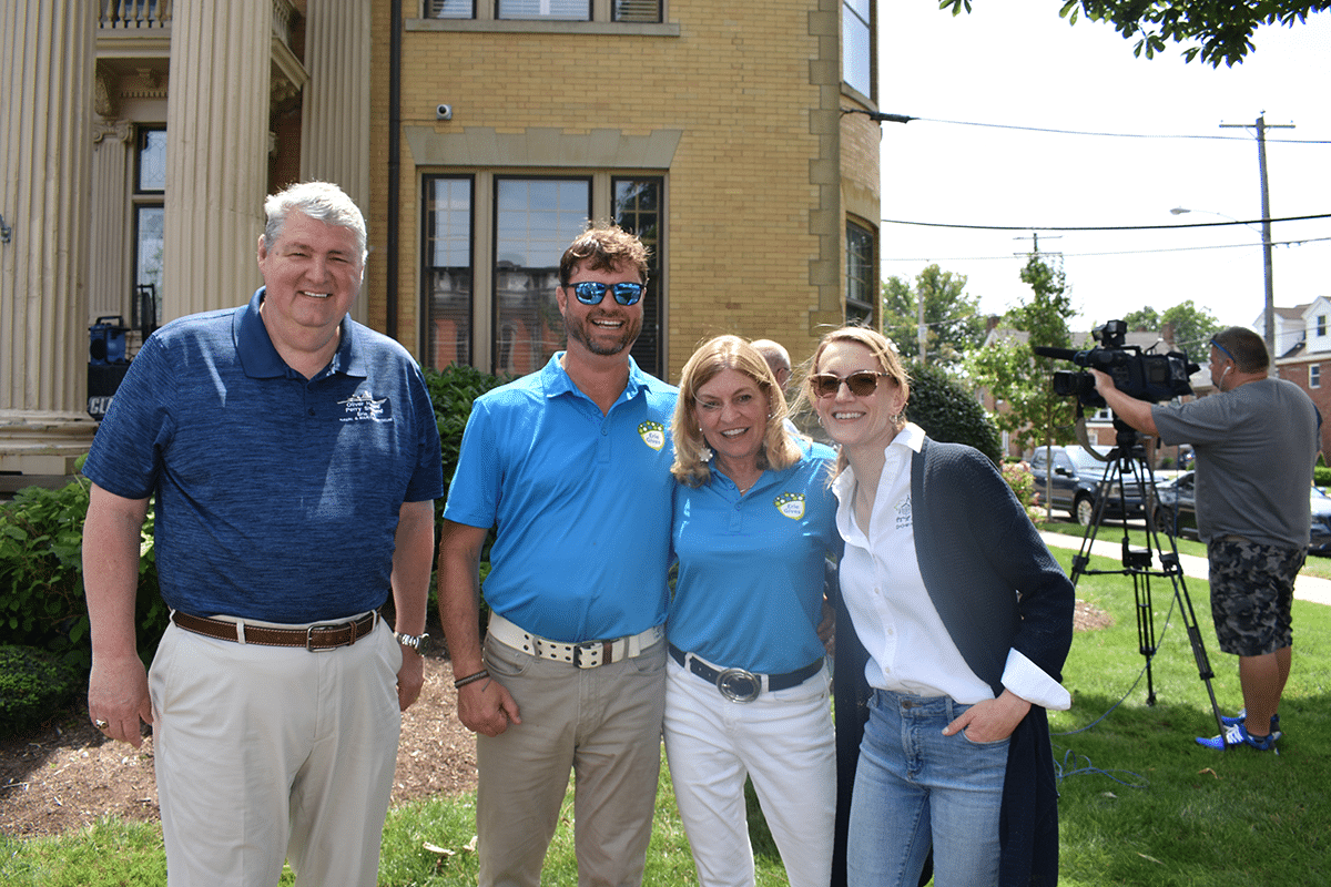 Four adults stand smiling outdoors in front of a yellow brick building, with a cameraman filming in the background on the right. Two people wear matching blue shirts and two are dressed casually. The weather appears sunny.