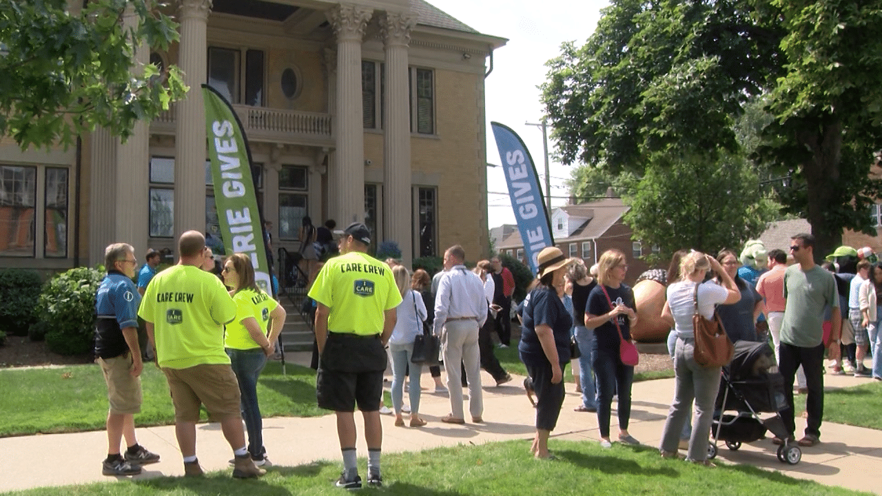 A group of people gather outside a large building with columns. Several wear bright yellow Care Crew shirts. Blue and green banners reading Erie Gives are visible among the crowd on a sunny day.