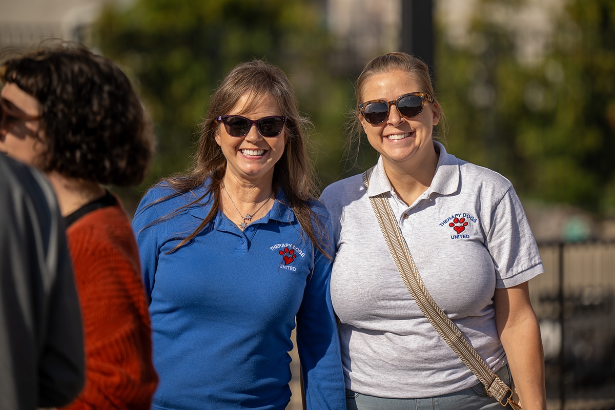 Two women wearing sunglasses and shirts with a red paw print logo and United text smile outdoors on a sunny day. One wears a blue shirt, the other a gray shirt with a crossbody bag. Other people are partially visible nearby.