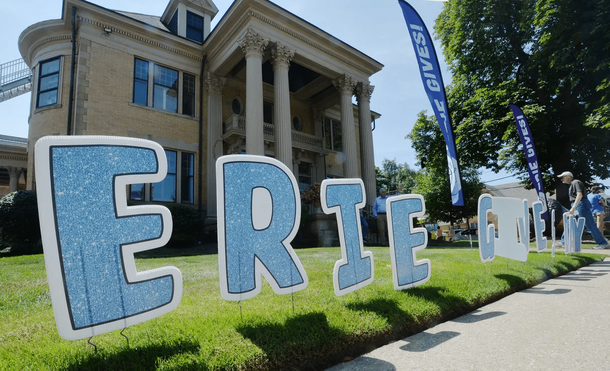 Large glittery letters spelling ERIE GIVES are displayed on a lawn in front of a historic building with columns on a sunny day. Blue flags in the background also read ERIE GIVES.
