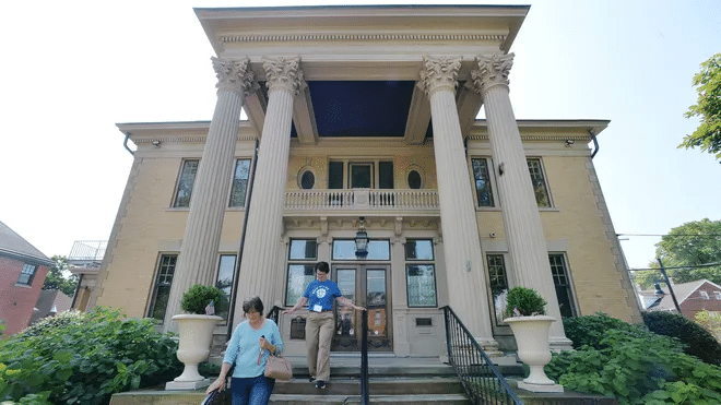 Two people walk down the steps of a grand building with tall columns, large windows, and decorative architectural details, surrounded by greenery on a sunny day.