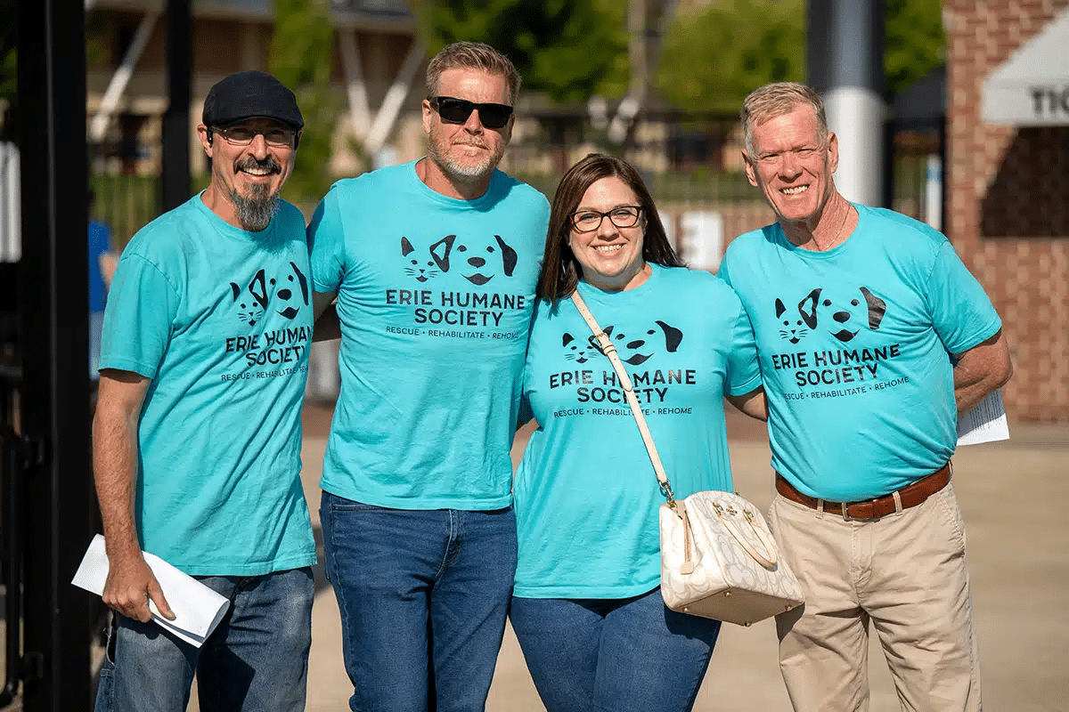 Four people wearing matching blue Erie Humane Society T-shirts stand together outdoors, smiling at the camera. Two hold papers, and one person carries a white purse. It appears to be a sunny day.