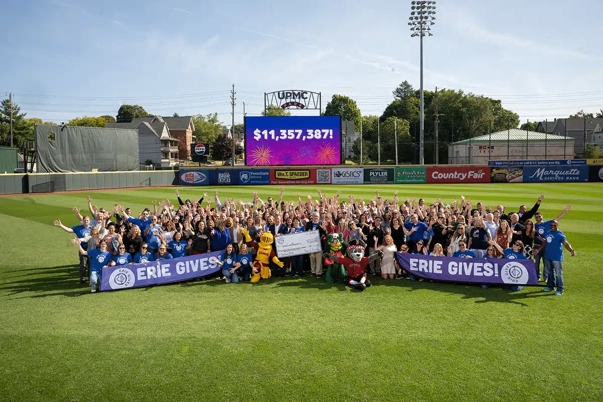 A large group of people stand on a baseball field holding ERIE GIVES! banners, cheering in front of a scoreboard displaying $11,357,387! and fireworks graphics. Several mascots are also present.
