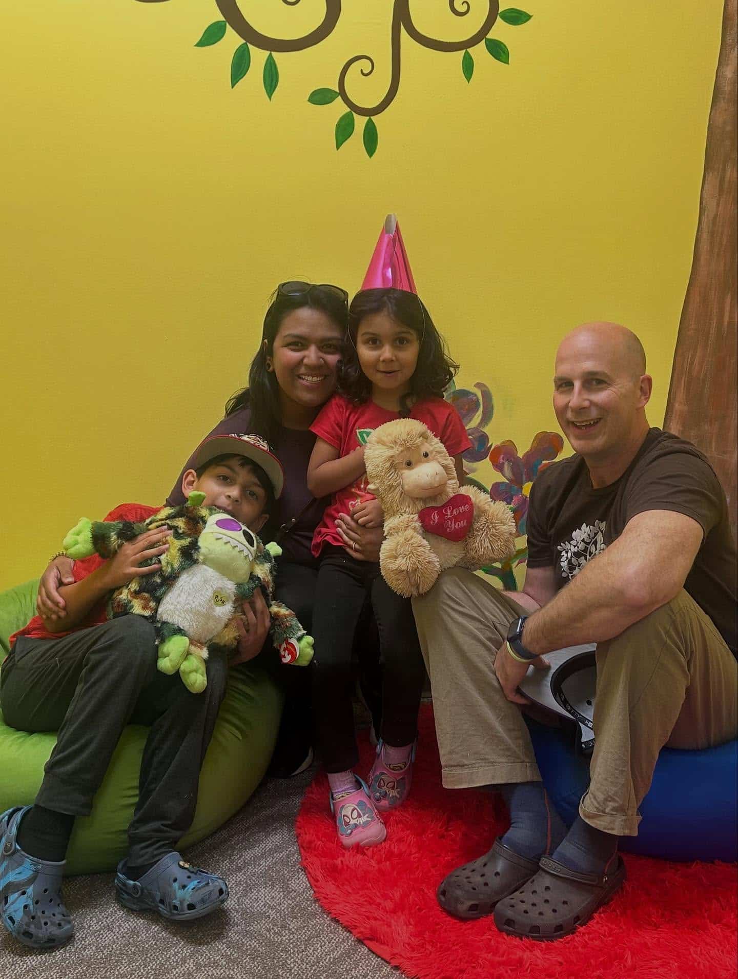 A woman and man sit with two children in a colorful room. One child wears a party hat and holds a teddy bear, while the other hugs a plush frog. Everyone is smiling and surrounded by bright decor and bean bags.