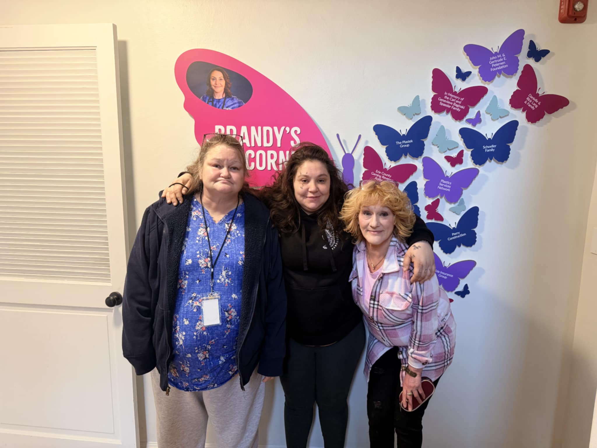 Three women stand together smiling in front of a wall display with colorful butterflies and the text Brandys Corner. The butterflies have messages on them, and one features a photo of a woman in a pink shape.