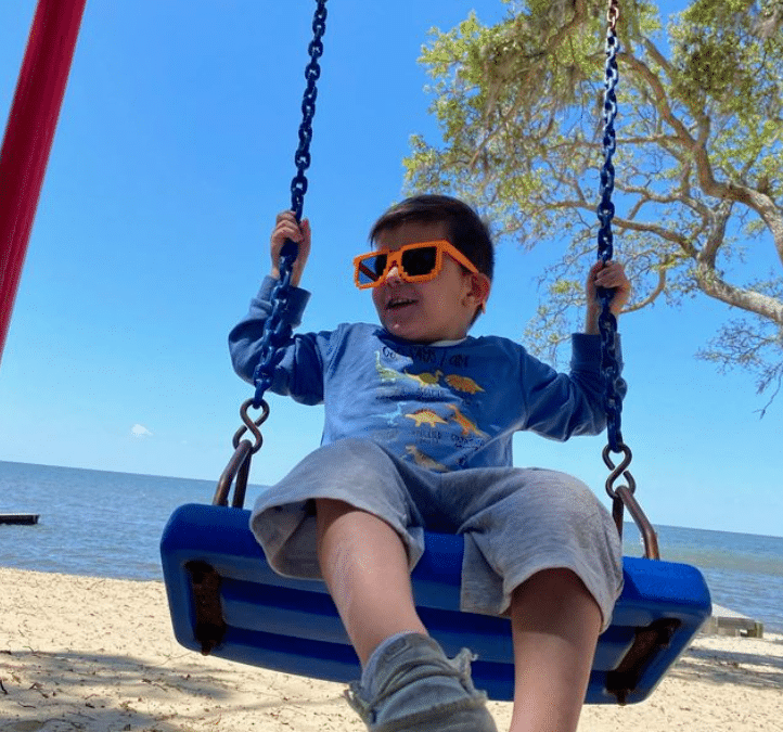 A child wearing orange sunglasses and a blue shirt with dinosaurs swings on a blue swing at a sandy beach, with water and trees in the background under a clear blue sky.