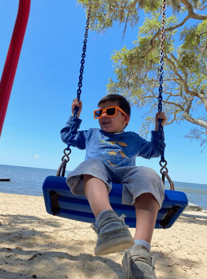A child wearing orange sunglasses and a blue shirt with dinosaurs swings on a blue swing at a sandy beach, with water and trees in the background under a clear blue sky.