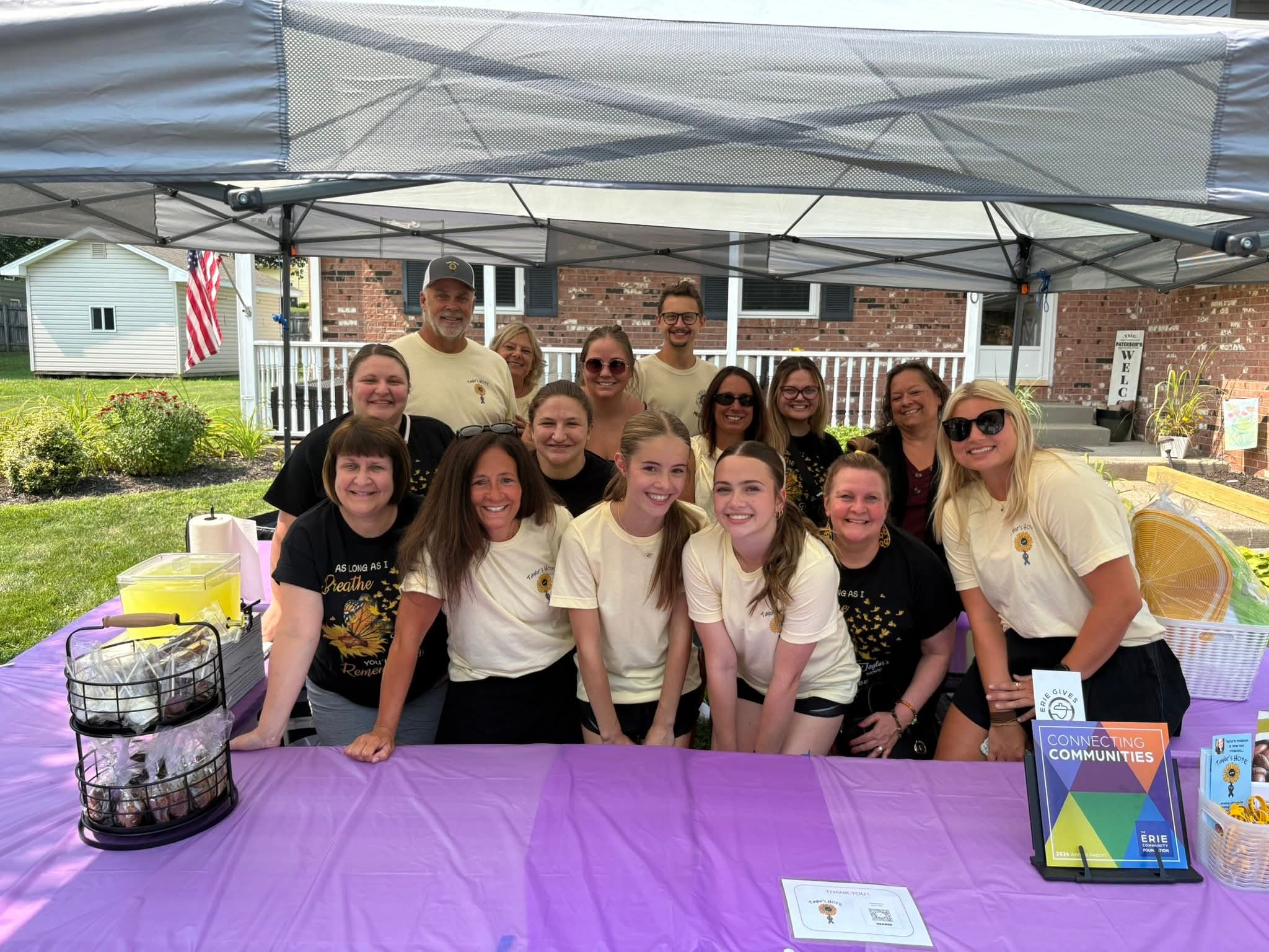 A group of people in matching t-shirts smile behind a table with a purple tablecloth at an outdoor event booth. Pamphlets, a drink dispenser, and a display rack are on the table. A house and lawn are visible behind them.