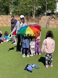 A group of young children and an adult gather under a small, colorful parachute on a grassy field, while other kids and an adult stand nearby. A jacket lies on the grass in the foreground.