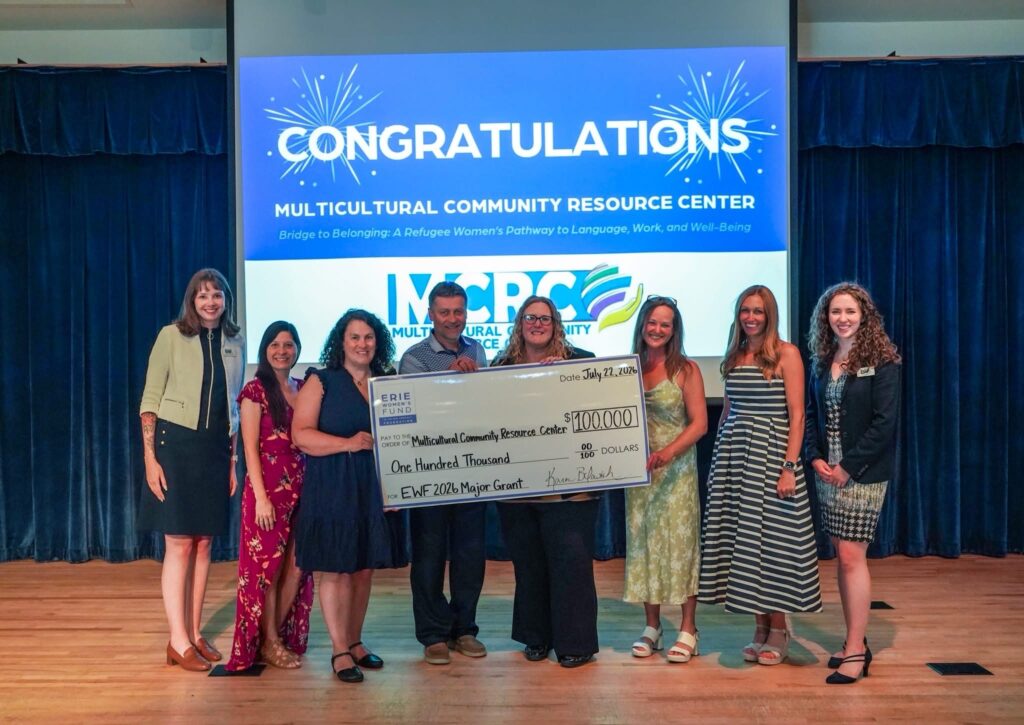 Eight people stand on stage holding a giant check for $100,000 from Erie Insurance, with a Congratulations sign for the Multicultural Community Resource Center displayed behind them.