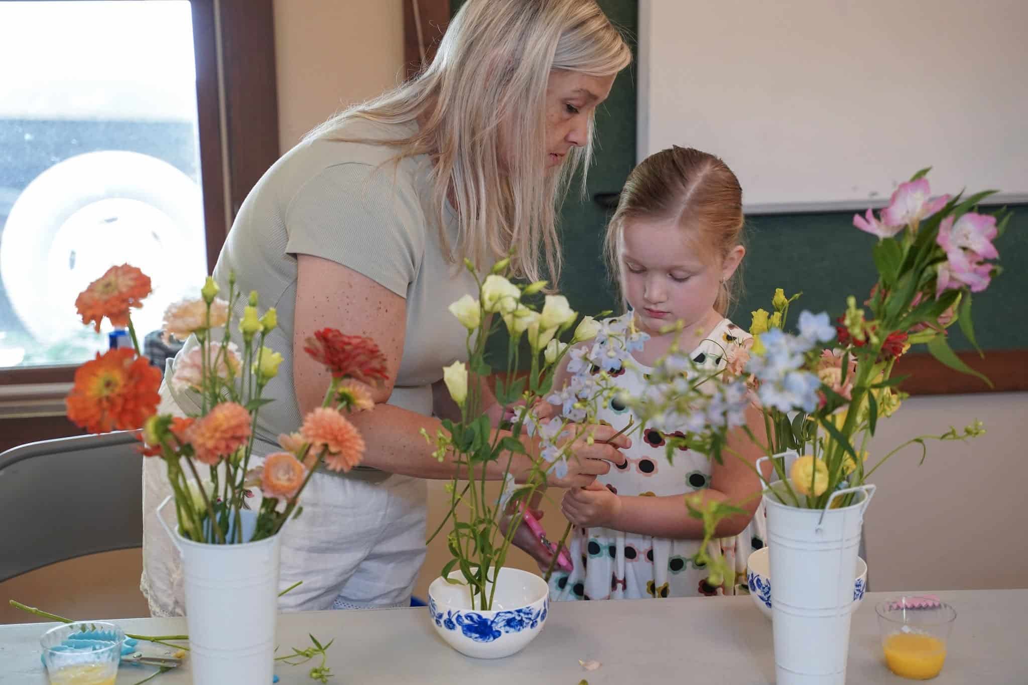 A woman helps a young girl arrange flowers in vases on a table. The girl wears a polka dot dress, and various colorful flowers are displayed. They appear focused on their activity inside a bright room.