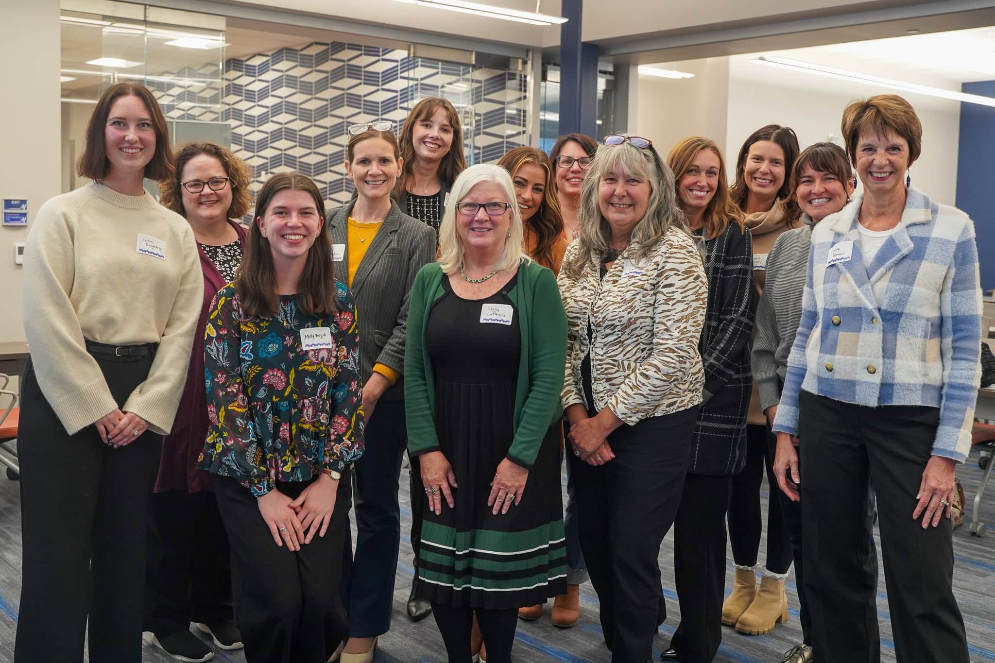 A group of thirteen women, standing together indoors, smiling at the camera. Most are wearing name tags, and they are dressed in business-casual attire. The background includes patterned walls and office furniture.