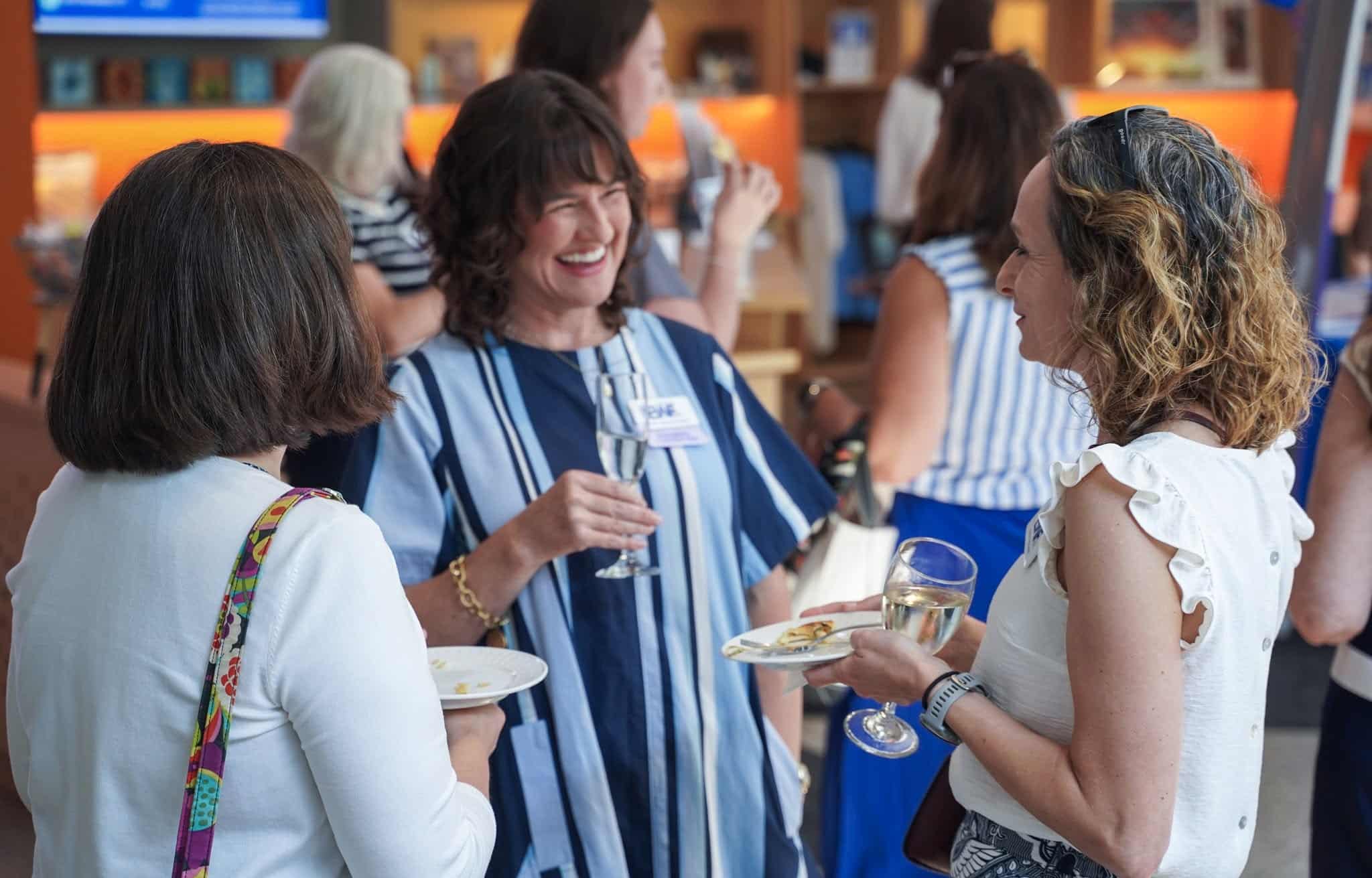 Three women stand together at a social event, smiling and talking while holding drinks and plates of food; other people are mingling in the background.