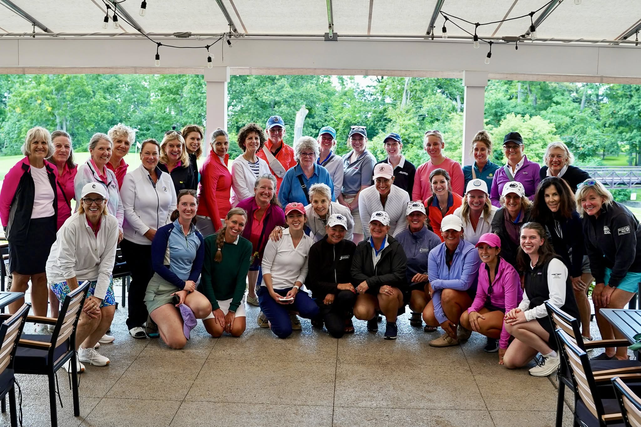 A large group of women in golf attire poses together under a covered patio, smiling at the camera. Outdoor greenery is visible in the background.