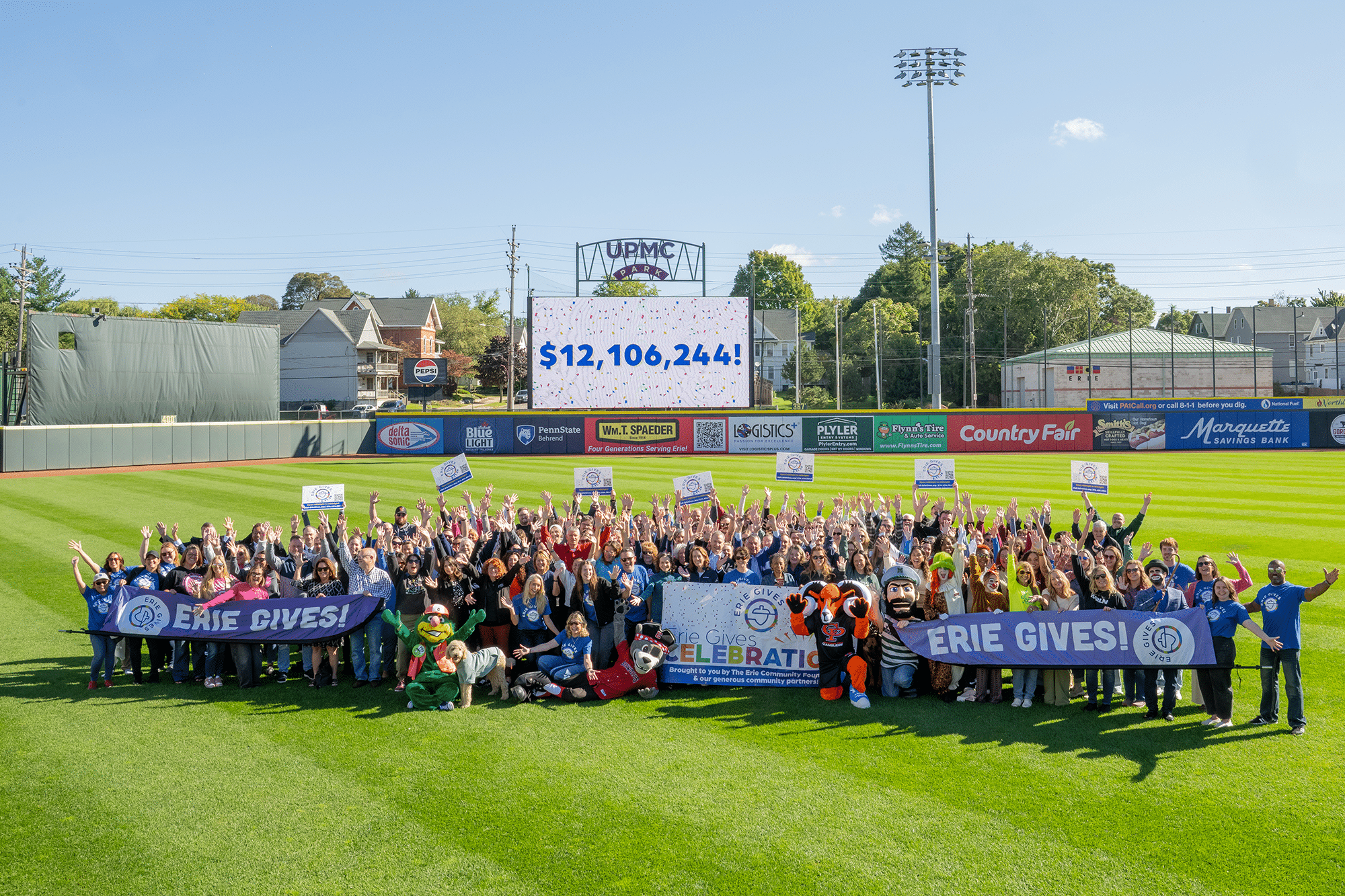 A large group of people on a baseball field holds Erie Gives! banners, celebrating in front of a scoreboard displaying $12,106,244! with trees, buildings, and advertisements in the background.