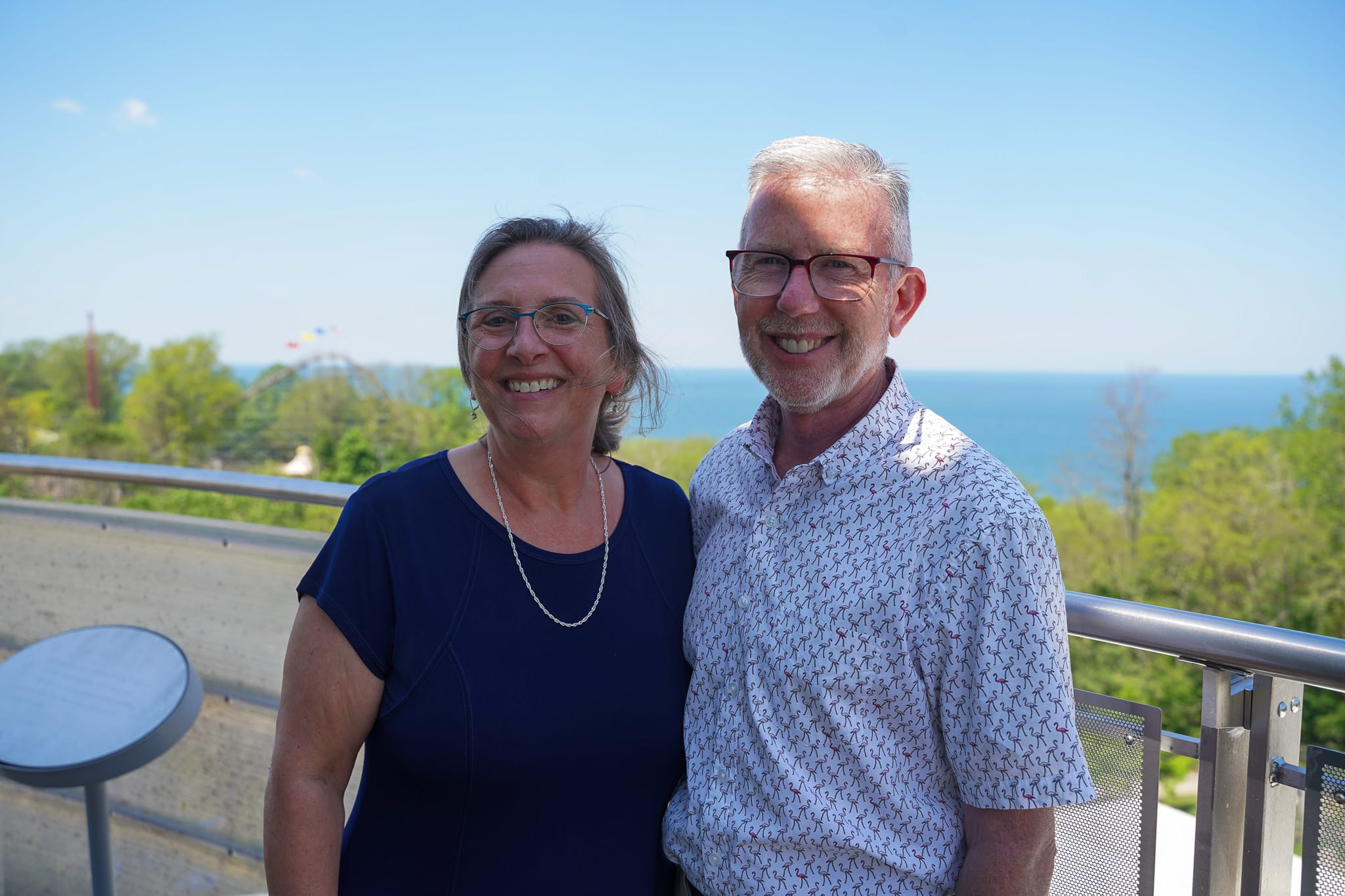 A smiling couple standing on an outdoor observation deck overlooking a lake and forested coastline.